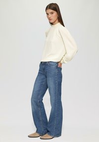 Blouse crème à manches longues avec un col froncé ; associée à un jean bleu évasé ; mannequin portant des ballerines beige, se tenant contre un fond clair.