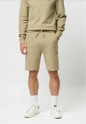 Mężczyzna w beżowych szortach i pasującej bluzie, białe skarpetki sportowe i białe trampki, stoi z rękami w kieszeniach.