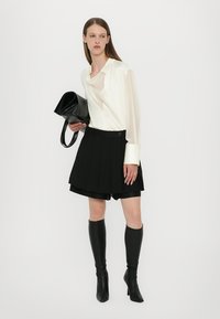 Pantaloni corti plissettati neri abbinati a una blusa bianca in seta. La modella porta una borsa in pelle nera e indossa stivali neri fino al ginocchio. Design semplice.