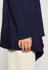 Pull marin avec une texture côtelée, des manches longues et un ourlet asymétrique. Présente un design ajusté avec une finition lisse sur la couche supérieure.