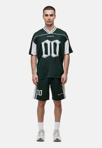 Ciemnozielony siatkowy t-shirt z białymi akcentami, ozdobiony dużą białą grafiką "00". W zestawie ze spodniami i białymi skarpetkami sportowymi.