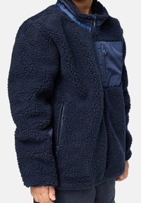 Personne portant une veste polaire bleu marine avec poches zippées sur la poitrine et sur les côtés, debout devant un fond uni.
