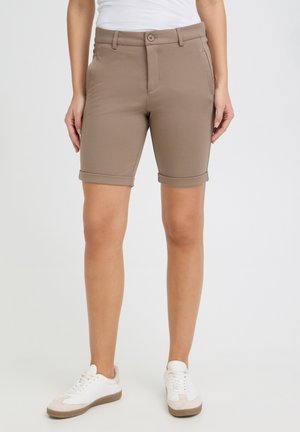 Naine, kes kannab kergeteääriseid beeže põlvekõrguseid lühikesi pükse, valgeid tossusid ja valget topi, seisab lihtsa heleda tausta ees.