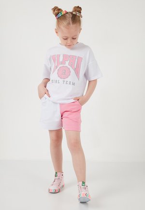 Ung pige med dobbelte knolde iført en hvid "Alpha Girl Team"-shirt, hvide og lyserøde shorts samt lyserøde høje sneakers med farverige såler.