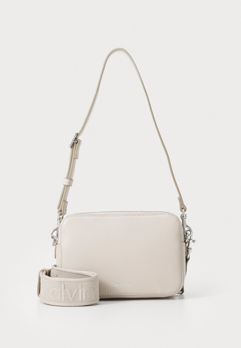 Lys beige crossover-veske med glatt skinn tekstur, sølvfarget hardware og en avtakbar stropp med preget logo. Rekteangulær form.