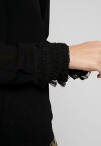 Svart topp med en genomskinlig, fransad manschett. Manschetten har lösa, texturerade trådar som skapar en lagrad look, vilket kontrasterar med det fasta tyget.