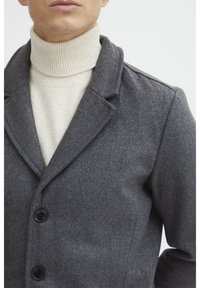 Manteau en mélange de laine gris avec un col à revers, trois boutons noirs, et un doux col roulé crème en dessous, présenté sur un fond uni.