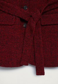 Veste à motif pied-de-poule rouge et noir avec des boutons noirs et une ceinture en tissu nouée à la taille.