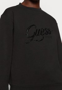 Czarny szary sweatshirt z połyskującym, czarnym logo "Guess" ozdobionym kryształkami oraz rokiem "1981" w kontrastowej fakturze. Miękki, strukturalny materiał.