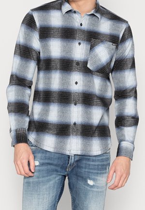 Homme portant une chemise à carreaux boutonnée à manches longues noire, bleue et blanche avec un jean bleu délavé, mains détendues le long du corps.