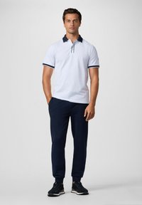 Biely polo tričko s námorníckym golierom a akcentmi na rukávoch, v kombinácii s námorníckymi teplákmi a čiernymi teniskami, s uvoľneným a priliehavým strihom.