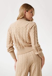 Beige-Kabelstrickpullover mit geripptem Kragen und Bündchen, der ein strukturiertes Muster und eine taillierte Passform aufweist; von hinten gezeigt.