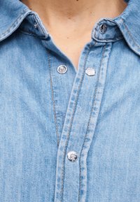 Ανοιχτό μπλε πουκάμισο denim με ασημένια κουμπιά snap, μερικώς ανοιχτό γιακά, εμφανές δέρμα στο σημείο του λαιμού.