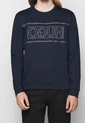 Persoon draagt een marineblauwe sweat met het woord "HUGO" in witte contouren achterstevoren gedrukt over de borst, met de tekst in kleine letters herhaald boven en onder.