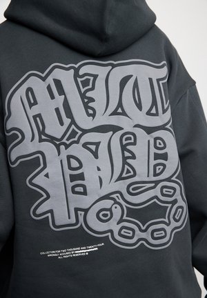 Person wearing black hoodie with large gray graffiti-style text and chain graphic on the back, small white printed text below.