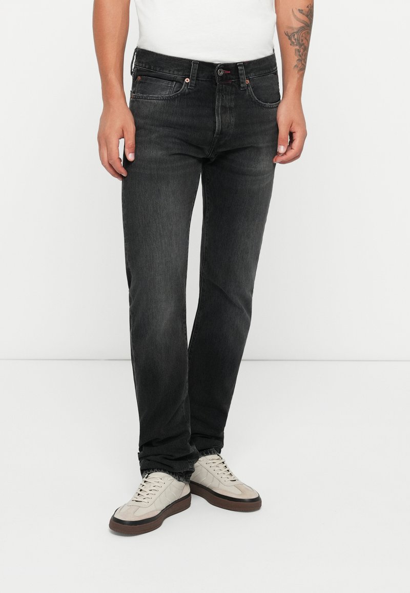Jeans en denim noir avec une coupe droite, cinq poches, des coutures beige clair et une fermeture à bouton, portés avec des baskets beige clair.