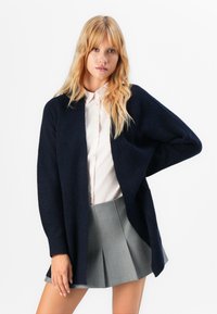 Cardigan ouvert en tricot marine avec des manches longues, en tissu texturé et coupe décontractée, associé à une chemise à boutons rose clair et une jupe plissée grise.