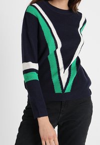 Granatowy sweter z ukośnymi zielonymi i białymi paskami, prążkowaną fakturą oraz luźnym krojem. Akcenty tworzą geometryczny wzór na przodzie.