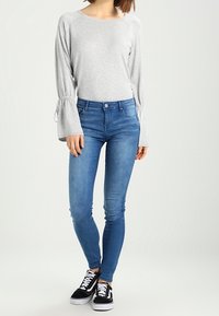 Šedý top s dlouhým rukávem a nařasenými detaily u manžet, doplněný modrými úzkými džíny. Outfit doplňují černobílé tenisky.