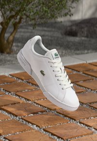 Sapatilha de couro branca com logótipo verde da Lacoste, bico redondo, sola texturizada e design de atacadores, exibida acima de uma superfície de azulejos padronizados.