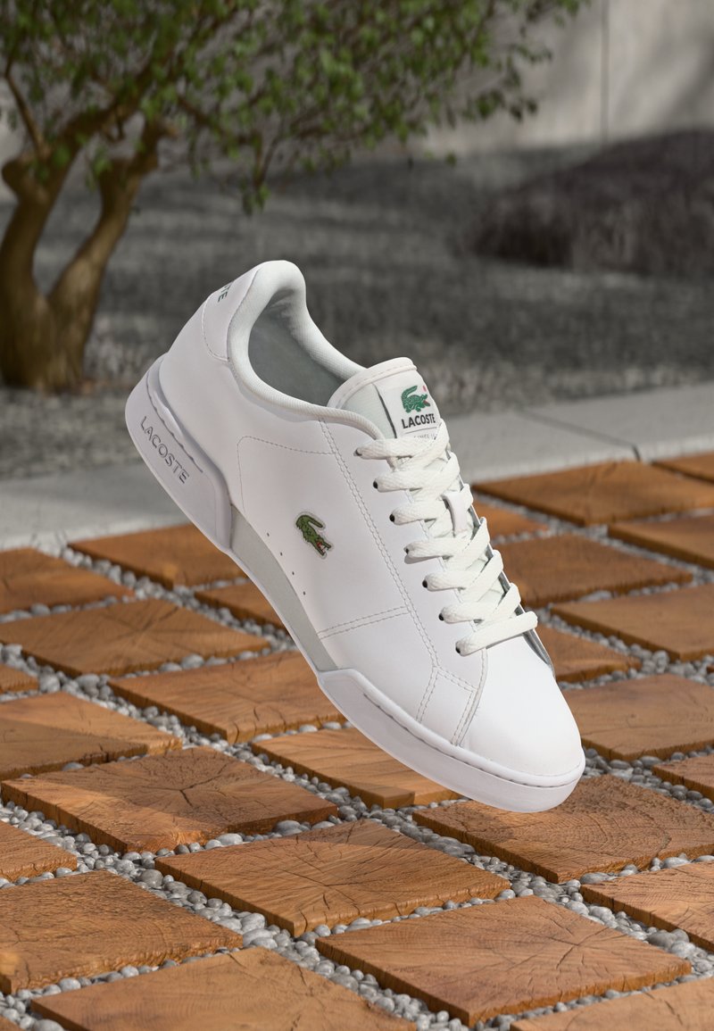 Sapatilha de couro branca com logótipo verde da Lacoste, bico redondo, sola texturizada e design de atacadores, exibida acima de uma superfície de azulejos padronizados.