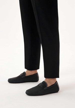 Beine in schwarzen, gekürzten Hosen und schwarzen Slip-on-Loafers mit strukturiertem Obermaterial und Lederbesatz, die auf einem weißen Boden stehen.