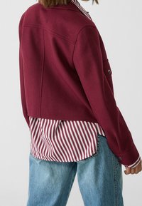 Burgunderfarvet cropped jakke lavet af uld, med en let tekstureret overflade, skuldersømme og en enkelt lomme. Sat sammen med en rød og hvid stribet skjorte.