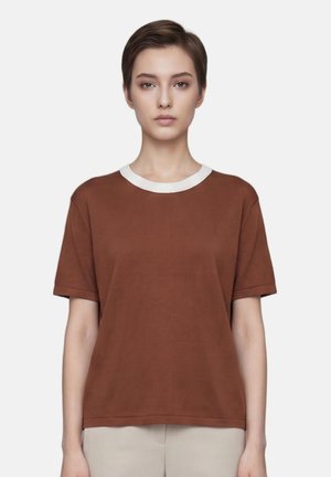 Jeune personne aux cheveux courts portant une chemise marron à manches courtes avec un col blanc, debout devant un fond uni.