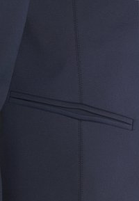 Detailní záběr na tmavě modrou látku s horizontální kapsou na lem a viditelným vertikálním švem.
