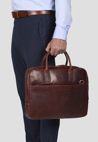Brun lærveske med doble håndtak, glidelås-lukking og frontlomme. Glatt tekstur med minimal merking og et stilrent design.