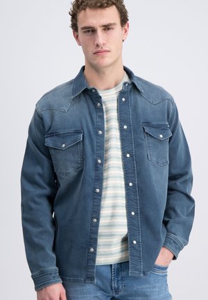 Junger Mann trägt ein aufgeknöpftes blaues Jeanshemd über einem hell gestreiften T-Shirt und Jeans, mit einer Hand in der Tasche, neutralem Gesichtsausdruck.