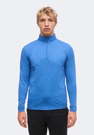 Jeune homme portant un haut bleu à manches longues zippé avec un motif de texte subtil répété et un pantalon noir, debout devant un fond uni.