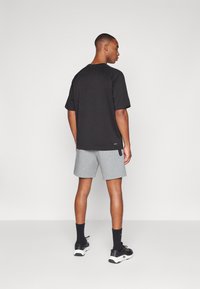 Uomo con maglietta nera a maniche corte, pantaloncini grigi, calzini neri e scarpe da ginnastica nere, in piedi di spalle su sfondo semplice.