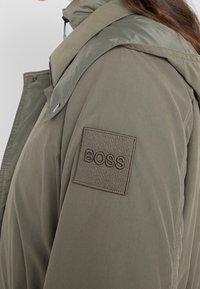 Olivgrön huva jacka med matt finish, med ett texturerat tyg, en framträdande BOSS-logotyp på ärmen och en dragkedja.