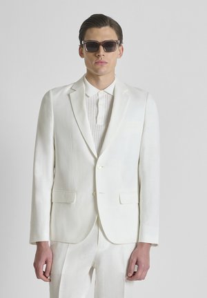 Hombre con traje blanco y jersey de punto con cuello tipo polo, de pie con las manos relajadas, lleva gafas de sol oscuras sobre un fondo gris liso.