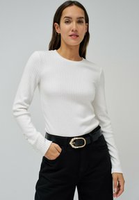Witte geribbelde langemouw top met een ronde halslijn, gecombineerd met zwarte broek en een zwarte riem met een gesp versierd met goud en parels.