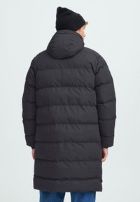 Černá péřová bunda s kapucí, s horizontálním prošíváním a volným střihem. Nositá přes modré džíny, texturovaná tkanina. Jednoduchý design.