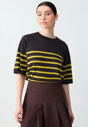 Jonge vrouw met donker haar in een gevlochten knot, gekleed in een zwart shirt met gele horizontale strepen en een donkerbruine geplooide rok.