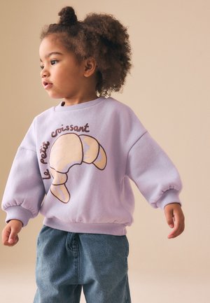 Småbarn med krøllet hår iført en lavendelfarvet sweatshirt med en croissantgrafik og tekst, parret med løstsiddende blå jeans, ser mod venstre.
