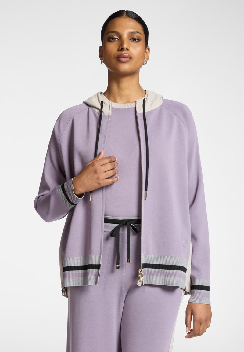 Giacca con zip color lilla con cappuccio bianco, cordini neri e dettagli a righe. Tessuto morbido, vestibilità rilassata, con dettaglio del logo all'orlo.