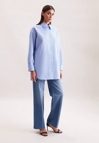 Gestreept blauw-wit shirt met lange mouwen en knopen aan de voorkant, gecombineerd met lichtblauwe wijdere spijkerbroek en zwarte sandalen met hak.