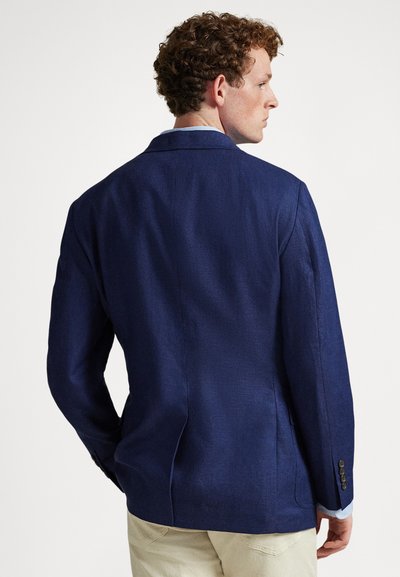 Mladý muž s kudrnatými vlasy otočený zády, oblečený v tmavě modrém saku přes světlou košili a béžové kalhoty na jednobarevném pozadí.