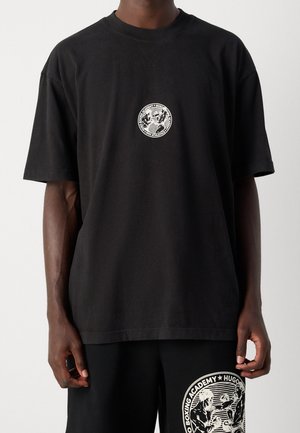 Mężczyzna ubrany w czarną, oversize'ową koszulkę i szorty z białym, okrągłym logo akademii boksu na piersi i lewym udzie.