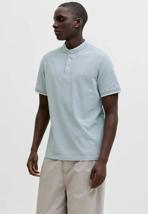 Młody mężczyzna o ciemnej karnacji, ubrany w jasnoniebieską koszulkę polo z krótkim rękawem i beżowe spodnie, stojący na tle jednolitego jasnego tła, patrzący w lewo.