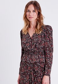 Vestito nero a fiori con maniche lunghe, vita smock e scollo a V; presenta motivi rossi e beige, texture del tessuto morbida.