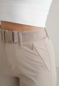 Béžové nohavice s hladkou textúrou, s širokým elastickým pásom so strieborným logom a malou zipsovou kapsou na boku.