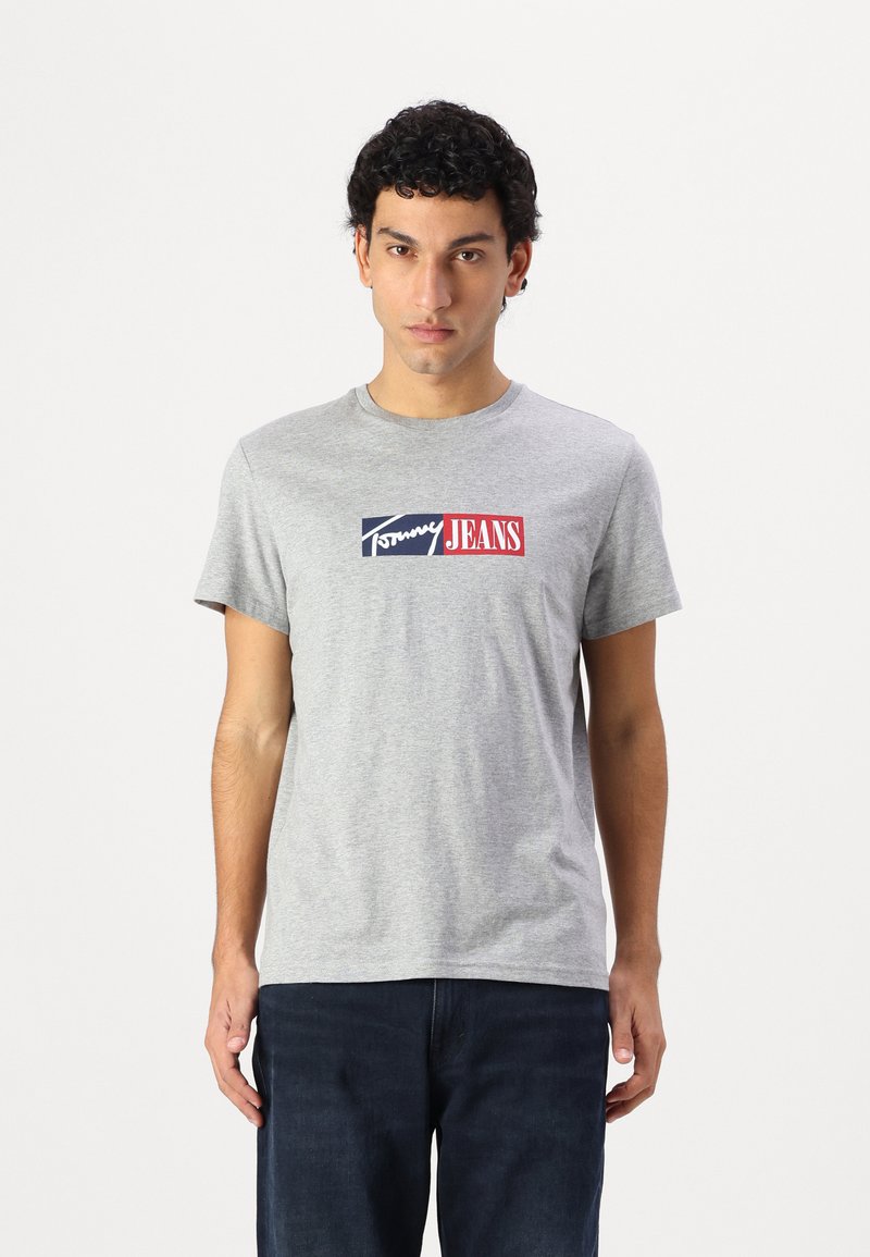 Pilkos spalvos medvilnės marškinėliai trumpomis rankovėmis, su stačiakampio formos logotipu mėlyna ir raudona spalvomis, kuriame yra užrašas "Tommy JEANS". Laidumo stilius, apvali iškirptė.