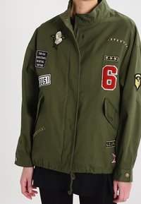 Oliwkowa kurtka z haftowanymi naszywkami, w tym biała kwiatowa, czerwona liczba 6 oraz szczegóły w stylu militarnym. Posiada zamek błyskawiczny i kieszenie.