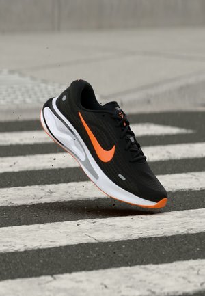 Chaussure de course Nike noire et orange en l'air au-dessus d'un passage piéton, avec de petits débris sous la semelle.
