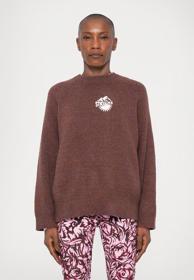 Brun, strikket sweater med rund hals og lange ærmer; har et hvidt grafisk logo på brystet. Sammen med rosa mønstrede leggings.